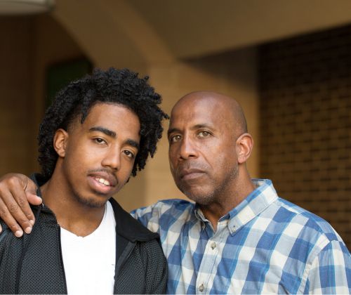 A father stands with his arm around his son's shoulder as they both look somber