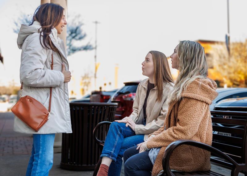 Outdoors scene of two women sitting on a bench and talking to another woman who is standing