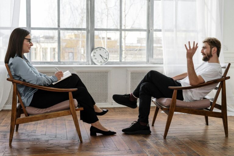 A man and woman sitting opposite each other, in a professional building. The woman is writing on a notepad while the man talks and gestures with his hand.