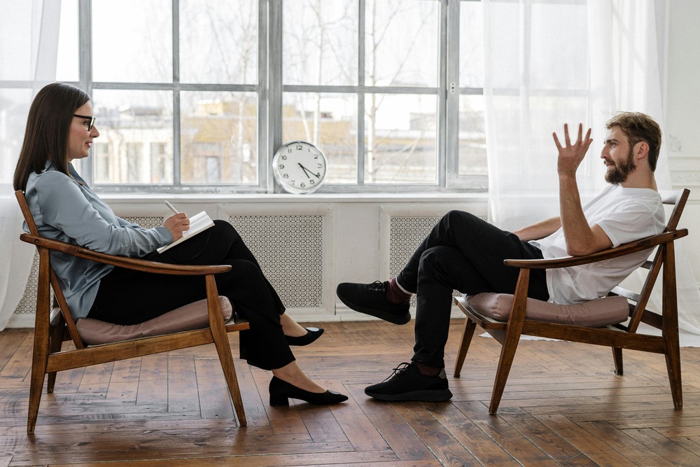 A man and woman sitting opposite each other, in a professional building. The woman is writing on a notepad while the man talks and gestures with his hand.