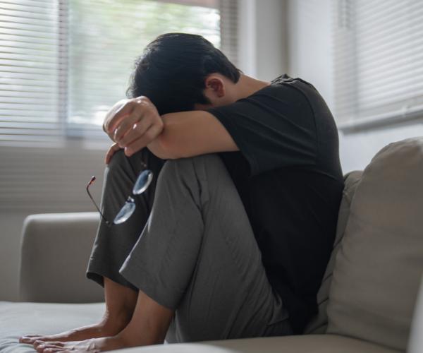 An anxious young man sits with his feet on a couch, head on his knees, and holding his glasses in one hand