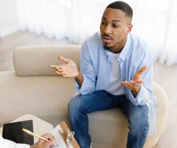 A young man talks to a partially visible therapist as she takes notes