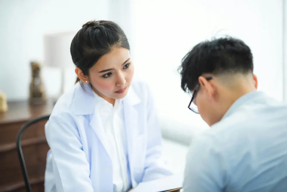 A young woman in a lab coat, looking concerned, sits across from a young man wearing glasses with his head down.