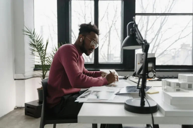 A young man sits at a desk in a white room, writing on a piece of paper.